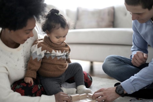 A young child in a brown sweater plays with wooden blocks, joined by a woman and a man